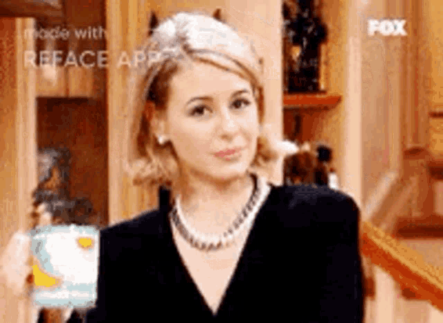 a woman in a black dress and pearl necklace is standing in front of a cake in a kitchen .