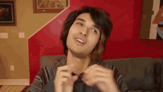 a young man with long hair is sitting on a couch in front of a red wall and making a funny face .