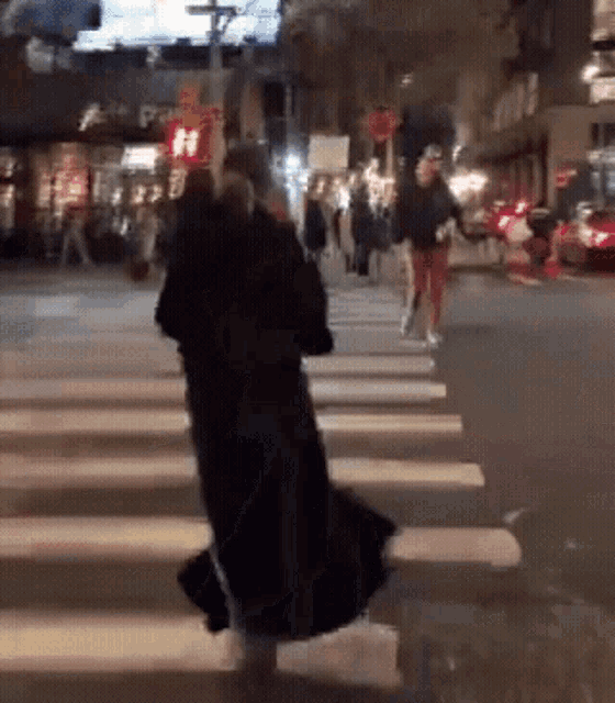 a woman in a black coat crosses a street at night