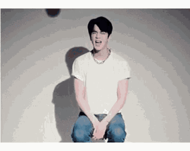a man in a white shirt and blue jeans is sitting on a stool and making a funny face .