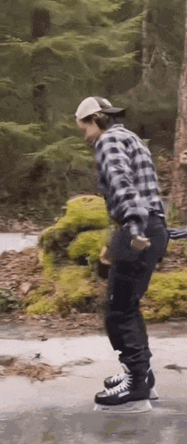 a man in a plaid shirt and black pants is riding a skateboard on a concrete surface in the woods .