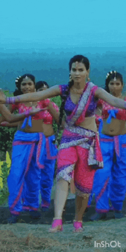 a woman in a pink and purple saree is dancing with a group of women in blue pants .