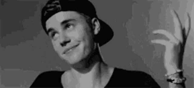 a black and white photo of justin bieber wearing a baseball cap and making a peace sign .
