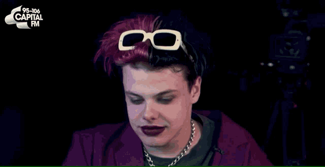 a man with red and black hair wearing glasses and a capital fm logo in the background