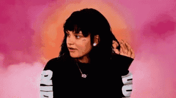 a woman is standing in front of a pink background . she is wearing a black shirt with the word ugg on the sleeves .