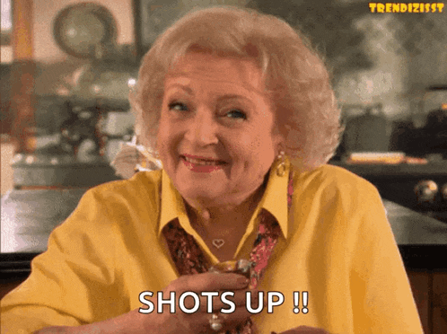 an older woman in a yellow shirt is holding a shot glass and says shots up