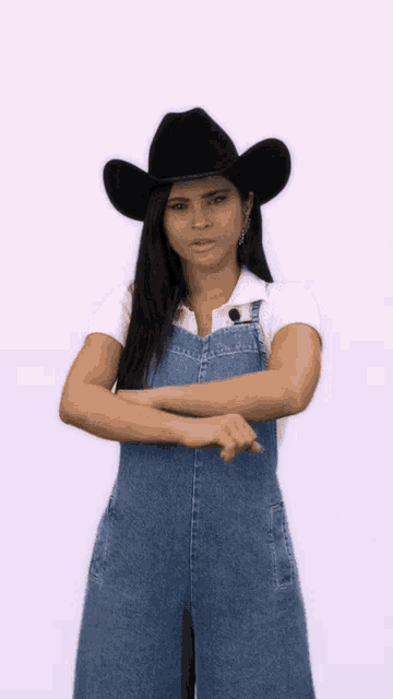 a woman wearing overalls and a cowboy hat is standing with her arms crossed