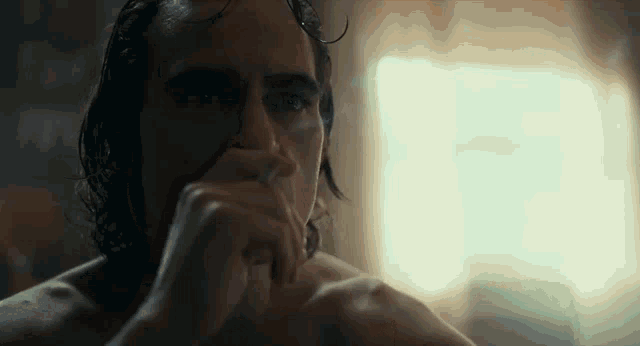 a man without a shirt is sitting in front of a window with his hand to his mouth .