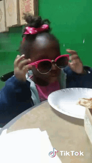 a little girl wearing sunglasses sits at a table eating pizza
