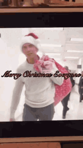 a man wearing a santa hat is carrying a pink bag and the words merry christmas everyone are on the screen