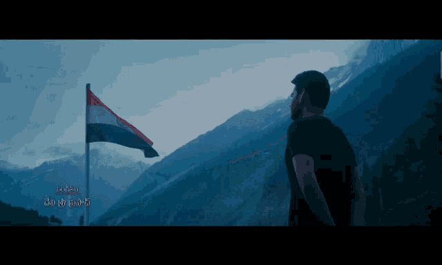 a man in a black shirt stands in front of a mountain with a flag flying in the background