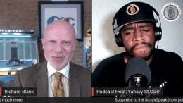 a man in a suit and tie is talking into a microphone while another man in a hat looks on .
