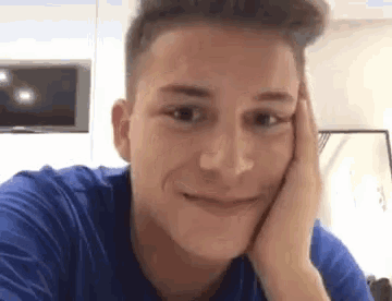 a young man in a blue shirt is holding his hand to his face and smiling .