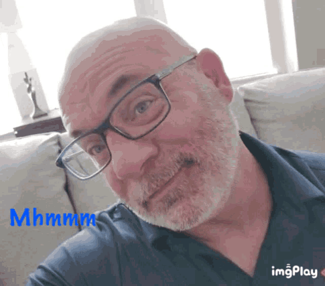 a bald man with glasses and a beard is sitting on a couch with the words mhmmm behind him