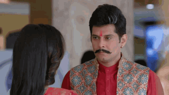 a man with a red bindi on his forehead talks to a woman