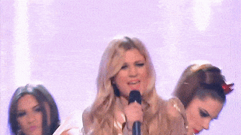 a blonde woman singing into a microphone on a stage with two other women behind her