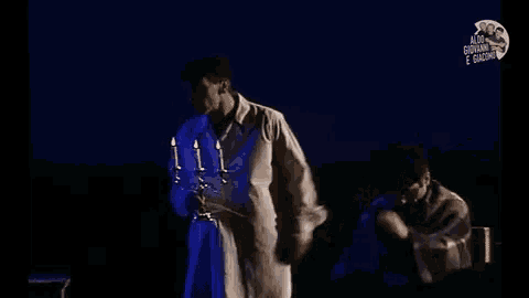 a man is standing on a stage holding a candle holder while another man looks on .