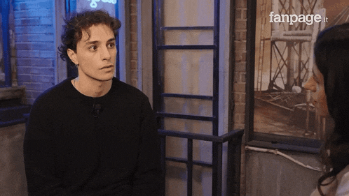 a man in a black sweater talks to a woman in front of a fanpage logo