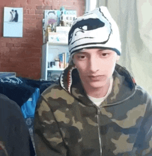 a young man wearing a camouflage hoodie and a beanie is sitting in a room .