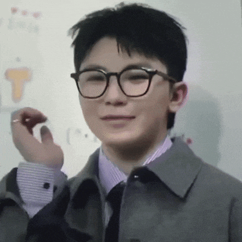 a young man wearing glasses and a jacket is standing in front of a whiteboard .