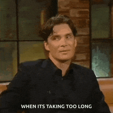 a man in a suit and black shirt is sitting in front of a brick wall and talking .