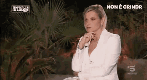a woman in a white suit is sitting at a table with her hands folded in front of a palm tree .