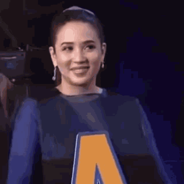 a woman in a blue dress is smiling and holding a large yellow letter a .