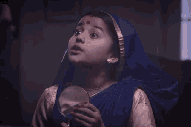 a little girl with a blue head scarf and a red dot on her forehead looks at herself in a mirror