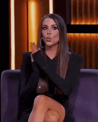a woman in a black dress is sitting on a couch with her legs crossed and making a funny face .