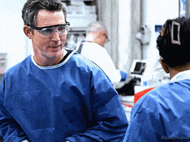 a man wearing goggles and a blue gown talks to a woman