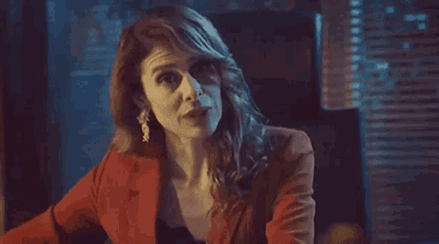 a woman in a red jacket and earrings is smiling while sitting in a dark room .