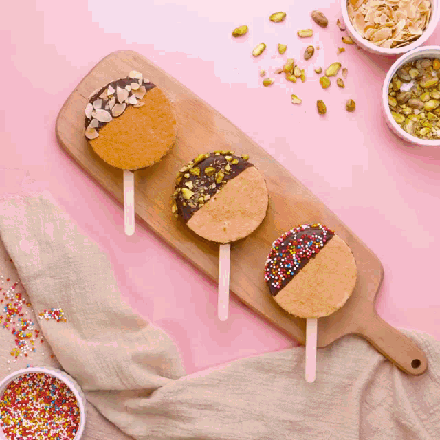 three cookies on a wooden cutting board with nuts and sprinkles on them