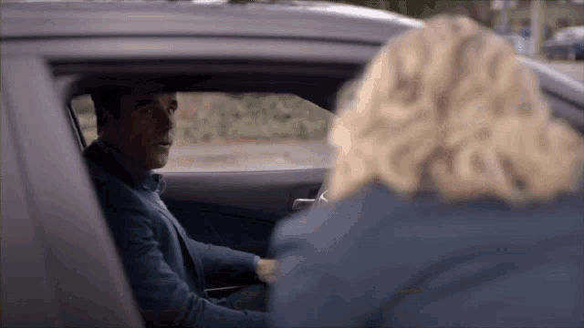 a man in a suit sits in a car talking to a woman in a blue jacket