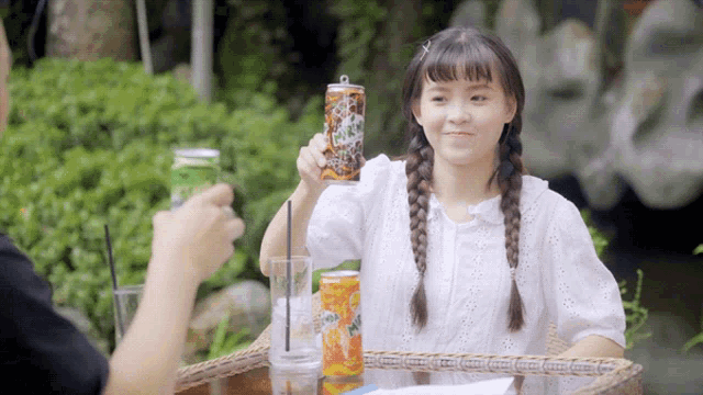 a girl sitting at a table holds up a can of soda