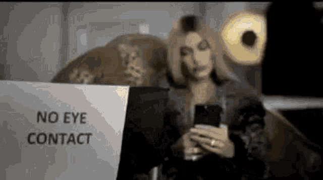 a woman is sitting in front of a sign that says `` no eye contact '' while holding a cell phone .