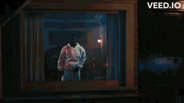 a man in a white hoodie stands in front of a window with a sign that says veed.io