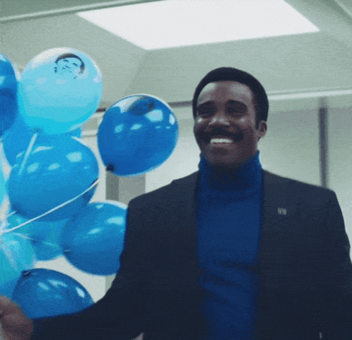 a man in a suit and turtleneck is holding a bunch of blue balloons and smiling