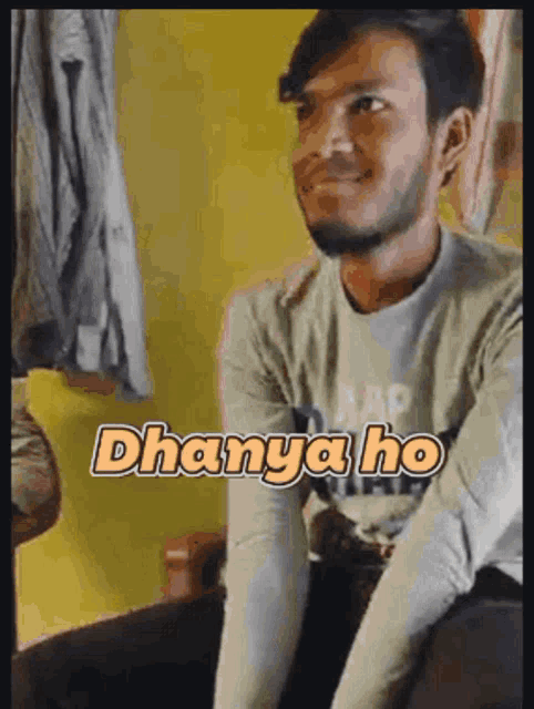 a man with a beard is sitting on a bed and smiling with the words dhanya ho written on his shirt .