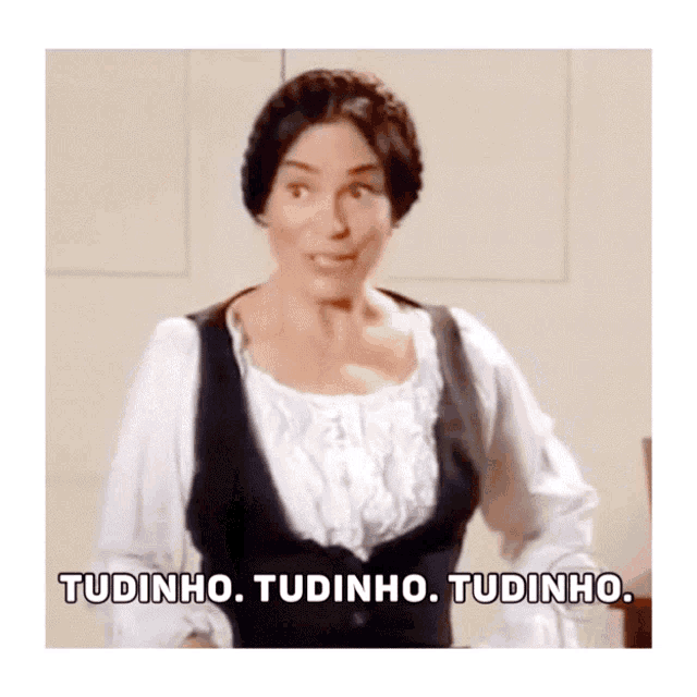 a woman in a white shirt and black vest says " tudinho tudinho tudinho "