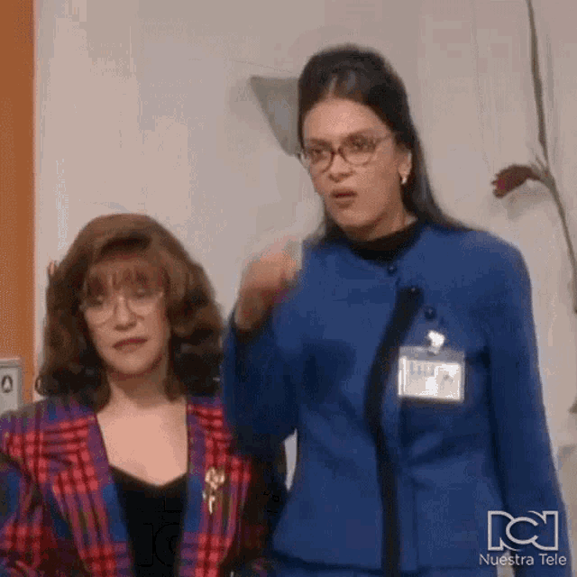 two women are standing next to each other in a room and one of them is wearing glasses .