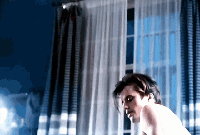 a man without a shirt is sitting in front of a window with blue curtains