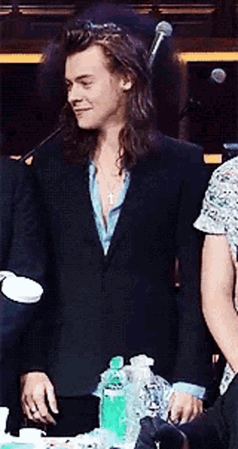 a man with long hair wearing a suit and a cross necklace is standing in front of a microphone .