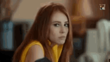 a woman with long red hair is wearing a yellow top and looking at the camera .