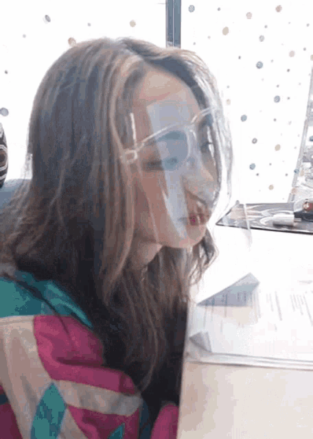 a woman wearing a face shield and glasses sits at a table