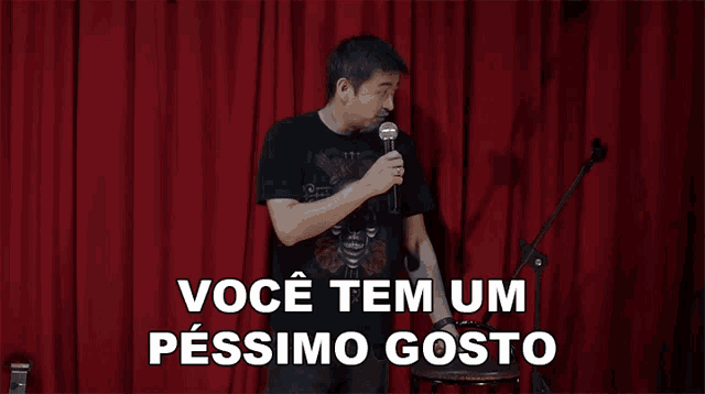 a man stands in front of a red curtain holding a microphone and says " você tem um pessim gosto "