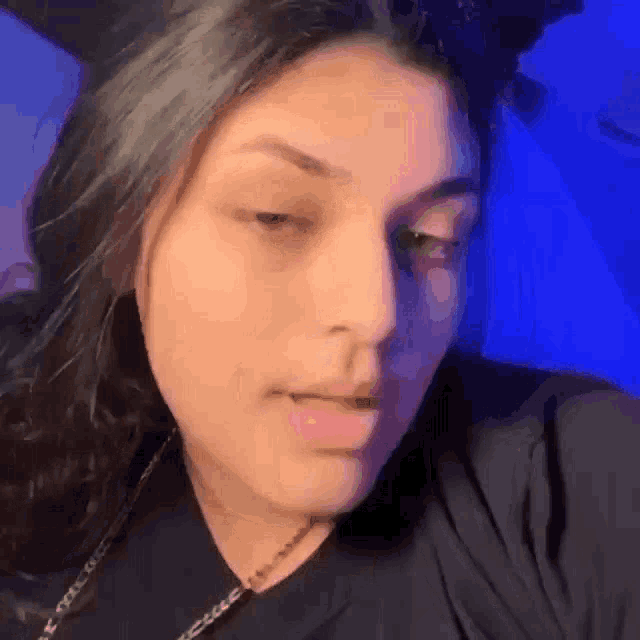 a close up of a woman 's face with a blue background . she is wearing a black shirt and a necklace .