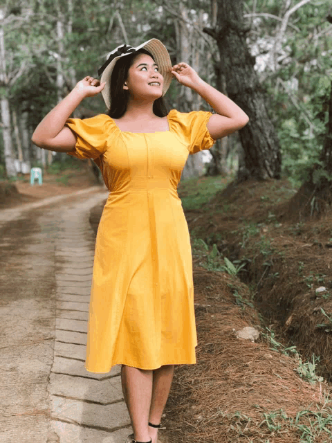 a woman wearing a yellow dress and a hat stands on a path