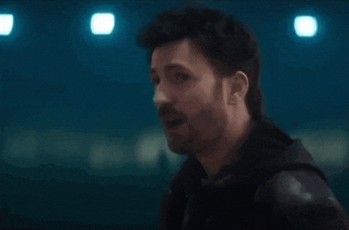 a man with a beard is standing in a dark room and talking to another man .