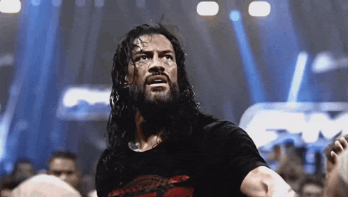 a wrestler with long hair and a beard is standing in front of a crowd and looking up .