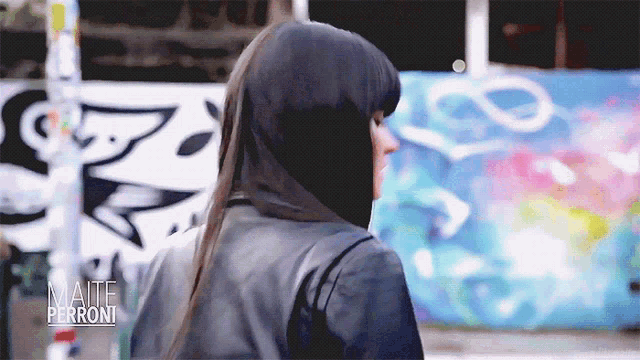 a woman in a black leather jacket is standing in front of a graffiti wall with the name maite perroni on the bottom